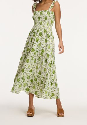 Femme portant une robe midi blanche sans manches avec imprimé floral vert et bretelles volantées, associée à des sandales compensées marron à bout ouvert.