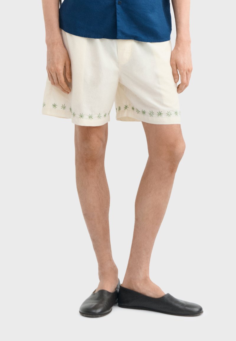 Personne portant un short crème avec des motifs d'étoiles vertes près de l'ourlet, une chemise bleue, et des chaussures noires sans lacets, debout les jambes croisées.