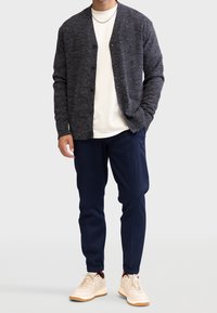 Szary, teksturowany kardigan z guzikami, noszony z białą koszulką i granatowymi spodniami, uzupełniony beżowymi trampkami. Prosty design, casualowy styl.