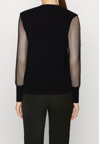 Maglione nero con maniche lunghe trasparenti, texture a costine nei polsini e nell'orlo. Indossato con pantaloni verdi a trama. Scollo a giro semplice sul retro.