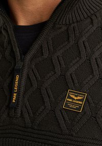 Pull-over en maille noire avec col zippé, présentant un motif texturé en losanges. Comprend une étiquette de marque avec des accents jaunes.