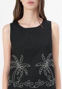 Femme portant un haut noir sans manches avec des palmiers blancs brodés et des volutes décoratives le long du bord inférieur.