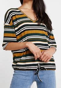 Top de cuello en V a rayas que presenta rayas horizontales en verde, mostaza, negro y blanco. Mangas cortas y un dobladillo con cordón para un ajuste ajustable.
