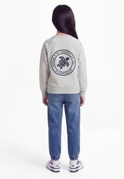 Kind mit langen dunklen Haaren, trägt einen hellgrauen Pullover mit Schildkröten-Logo und französischem Text auf der Rückseite, blaue Jeans und weiße Turnschuhe, schaut nach hinten.