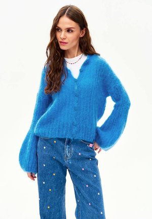 Kobieta ubrana w miękki, niebieski kardigan zapinany na guziki, na biały top, w połączeniu z niebieskimi dżinsami ozdobionymi małymi, kolorowymi haftowanymi kwiatami.