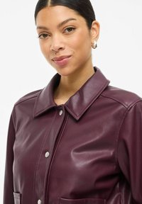 Veste en cuir bordeaux avec un col, des boutons-pression et deux poches avant. Texture lisse et design ajusté.