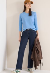 Femme portant une casquette marron, un haut bleu clair à manches trois-quarts, un jean bleu foncé, des baskets blanches, tenant une veste marron, debout contre un mur beige.