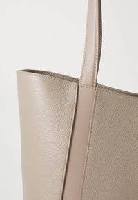Beige leather tote bag with a textured surface, vertical seam details, and slender, stitched shoulder strap. Smooth finish on edges.