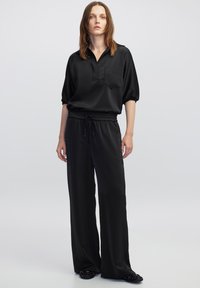 Zwarte, gladde stof bovenkant met een kraag en een zak, gecombineerd met losse zwarte broek met een elastische tailleband en trekkoord.