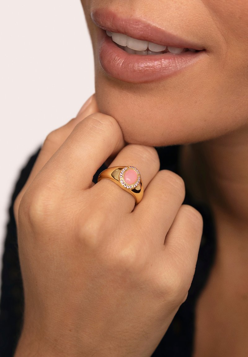 Anillo de oro con una piedra central rosa, rodeada de pequeñas piedras, llevado en una mano que descansa suavemente sobre la barbilla. Textura suave.