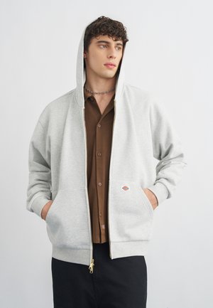 Ung mann iført lys grå zip-up hettegenser over brun skjorte med knapper, med hendene i lommer, ser sidelengs mot en ensfarget bakgrunn.