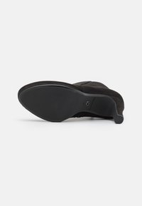 Chaussure à talon haut en daim noir posée sur le côté, montrant la semelle lisse et le talon bas sur fond blanc.