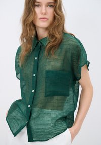 Camicia verde trasparente con bottoni, con un motivo testurizzato, maniche corte e una tasca applicata. Presenta una leggera trasparenza e una vestibilità comoda.