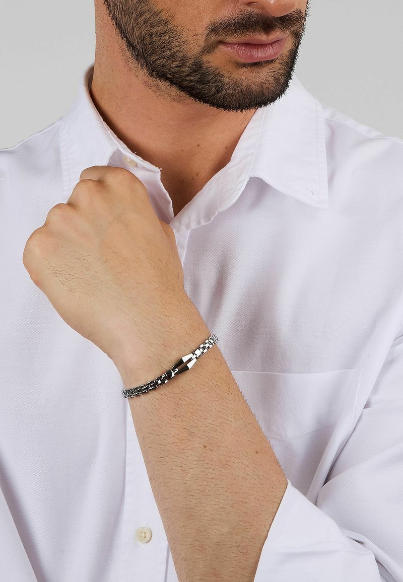 Bracciale metallico con maglie alternate; colore argento lucido con chiusura a gancio. Indossato su un polso ricoperto da peli corti e scuri.