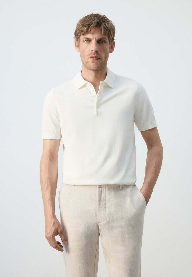 Homme debout, les mains dans les poches, portant un polo blanc à manches courtes et un pantalon beige sur un fond clair uni.