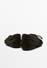 Tmavě hnědé semišové mokasíny s jednoduchým designem, s ozdobou ve formě uvázaného šňůrky a plochou gumovou podrážkou. Pohled zezadu ukazuje měkkou texturu.