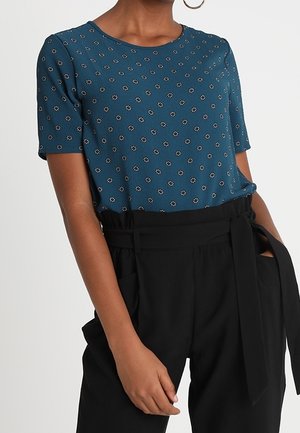 Vrouw draagt een teal kortemouwen top met kleine bloemmotieven en een zwarte broek met hoge taille en een gestrikte riem.