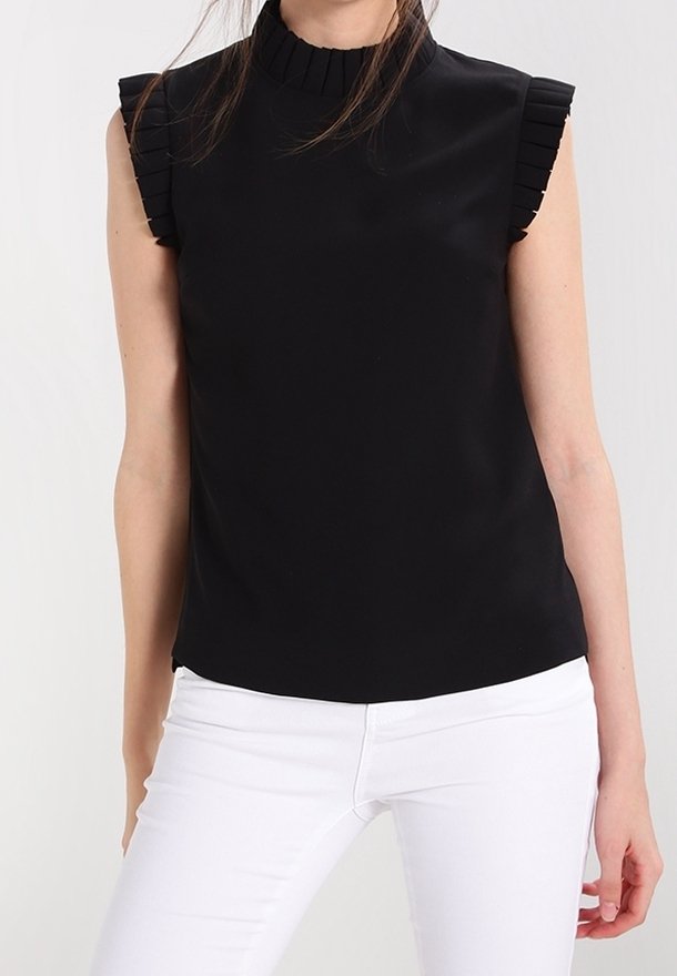 Haut noir sans manches avec un col haut et des accents froncés aux épaules, fabriqué en tissu lisse. Associé à un pantalon blanc.
