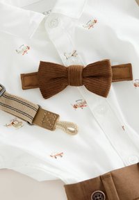White collared shirt with printed airplanes and vehicles, paired with a brown corduroy bow tie and suspenders featuring a tan strap and woven detail.