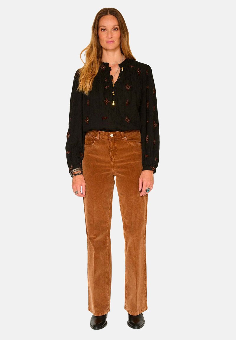 Blouse noire à manches longues avec des détails brodés et des boutons, associée à un pantalon en velours côtelé marron évasé et des bottines noires.
