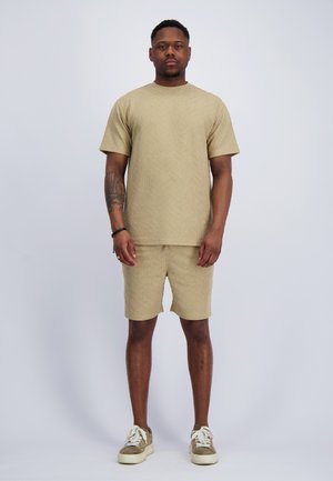 Mann steht, trägt ein beiges, strukturiertes kurzärmeliges Hemd und Shorts mit beigen Sneakers vor einfarbigem Hintergrund.
