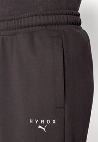 Schwarze Sporthose mit elastischem Bund, Seitentasche und weißem "HYROX"-Logo sowie einem kleinen weißen Puma-Logo. Glatte Textur.