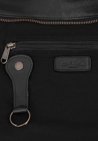 Close-up of black bag fabric with two horizontal zippers, a leather key holder with metal ring, and a stitched brand label.
