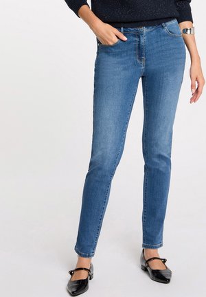 Donna che indossa jeans blu slim fit, maglione nero, scarpe nere modello Mary Jane e bracciale argento, in piedi con una mano in tasca.