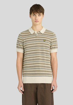 Giovane uomo con capelli corti che indossa una polo beige e marrone a maniche corte con fantasia e pantaloni marrone scuro, in piedi davanti a uno sfondo semplice.