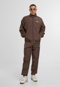 Brązowy dres wykonany z lekkiego materiału, składający się z kurtki na zamek z wysokim kołnierzem i elastycznymi mankietami, w połączeniu z zwężającymi się spodniami.