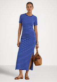 Žena v modro-bielych pruhovaných maxi šatách s krátkym rukávom a bočným rozparkom, drží hnedú koženú kabelku a má na nohách hnedé sandále.