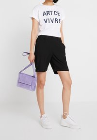 Donna che indossa una t-shirt bianca "ART DE VIVRE", pantaloni corti neri arrotolati, sneakers bianche, mentre tiene una piccola borsa viola con un cinturino da polso.