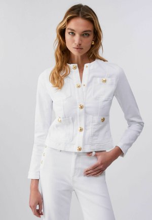 Femme aux longs cheveux ondulés portant une veste blanche avec des boutons dorés et un pantalon taille haute assorti, debout devant un fond uni.