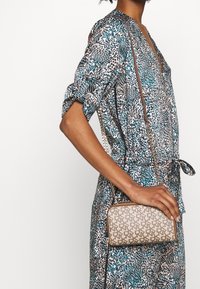 Beżowa torebka o fakturowanej powierzchni z geometrycznym wzorem, wyposażona w złoty łańcuszkowy pasek. W tle widoczny jest wielokolorowy strój w panterkę.