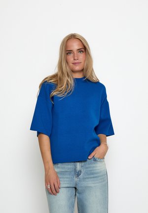 Blond kvinde med løst hår iført en klar blå kortærmet top og lyseblå jeans, stående med den ene hånd i lommen.