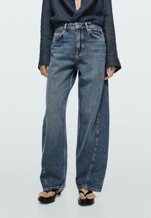 Osoba nosząca luźne niebieskie dżinsy z denimu, czarną koszulę z długim rękawem oraz czarne sandały na platformie z otwartym palcem.