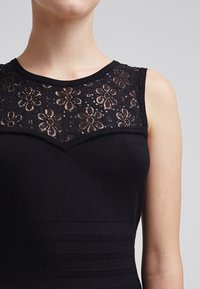 Abito nero con corpetto in pizzo floreale, impreziosito da paillettes. La parte inferiore presenta righe di texture e bande decorate. Design senza maniche.