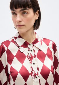 Femme aux cheveux courts et foncés portant un chemisier en satin à motifs losanges rouges et blancs, avec bordure volantée et boutons noirs.