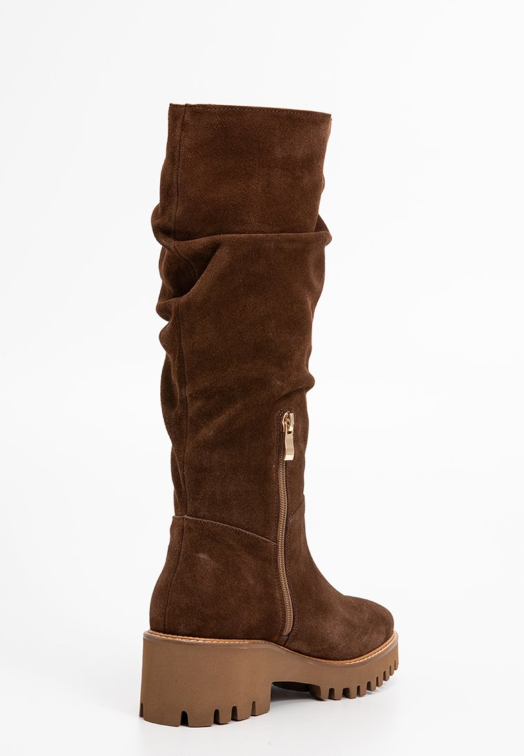 Bruine suède kniehoge laars met chunky rubberen zool, zijrits en slouchy bovendesign.