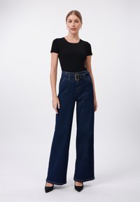 Sieviete, kas ģērbusies melnā īsām piedurknēm augstās jostasvietas platās kājās tumši zilās džinsās ar lielu jostu, stāv uz baltas fona.