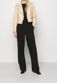 Beige lädermotorcykeljacka med silverdetaljer, svart topp och löst sittande svarta byxor; modellen bär svarta skor.