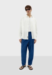 Witte knoop-shirt, met een gestructureerde stof, gedragen over een witte tanktop. Blauwe gestructureerde broek en zwarte teenslippers maken de outfit compleet.