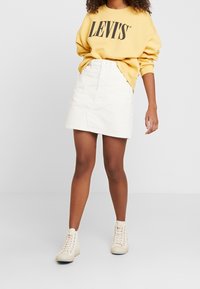 Felpa giallo chiaro con logo "LEVI'S", abbinata a una minigonna in velluto a coste bianca e stivaletti beige con lacci. Outfit semplice e casual.