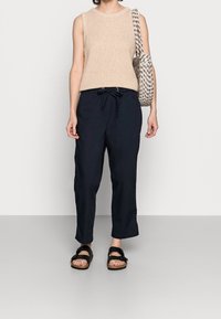 Kvinna i ärmlös beige stickad topp, svarta byxor med dragsko, svarta sandaler och bär en axelväska med svart-vitt mönster.