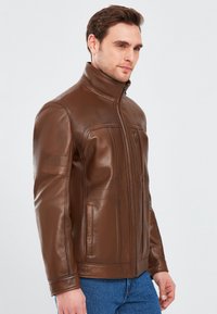 Giacca in pelle marrone con colletto alto, zip frontale e cuciture strutturate. Presenta due tasche laterali e una superficie liscia.