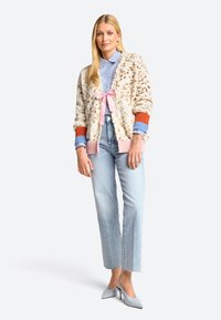 Cremfarbener, flauschiger Cardigan mit orangefarbenen, rosafarbenen und blauen Akzenten; hellblaues gestreiftes Hemd mit einer rosa Schleife; helle Jeans; graue spitze Absätze.