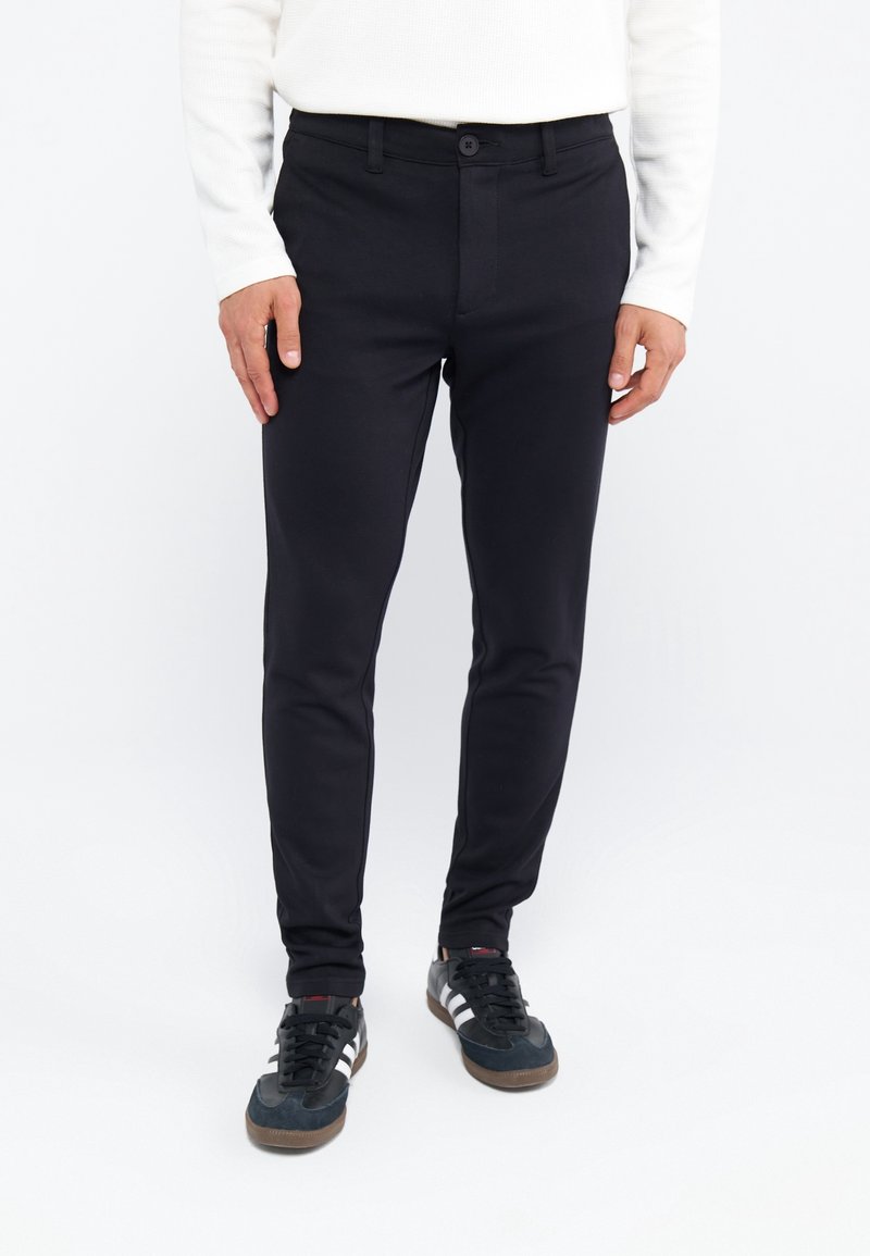 Zwarte slim-fit chinos met een gladde textuur, voorzien van een knoopsluiting en zijzakken, gecombineerd met zwart en marineblauwe sportschoenen.