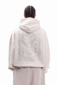 Sweat à capuche crème surdimensionné avec une capuche à cordon, présentant un graphique argenté détaillé dans le dos représentant une figure ailée accompagnée d'oiseaux.