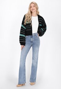 Črna skrajšana kardigan z teal in belo črtasto vzorcem, v kombinaciji z visokimi, svetlo modrimi razširjenimi kavbojkami in bež gležnjački.