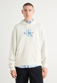 Sudadera con capucha color crema y un bolsillo frontal. Presenta un logo azul "CK" y el texto "calvin.klein". Tejido suave y corte relajado.